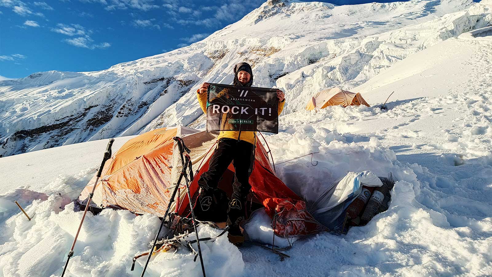 Bergsteiger steht vor Zelt im Schnee auf Manaslu und hält Fahne hoch mit der Aufschrift "Rock it"